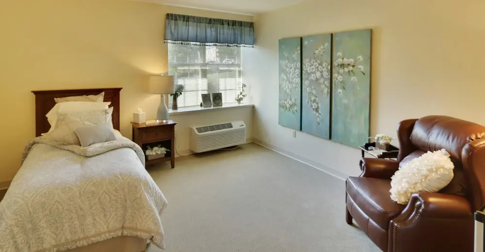 A bedroom with a twin bed, leather chair and floral painting at American House retirement home in West Knoxville